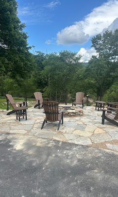 Irregular flagstone patio with a round stone fire pit, Adirondack chairs, and wooded green trees under a bright partly cloudy sky.