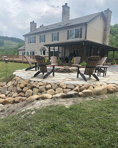Elevated circular flagstone patio with a river-rock retaining wall, Adirondack chairs around a fire pit, and a mountain home in the background.