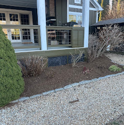 Mulched beds with stone edging and a gravel walk beside a home porch with green siding and white trim.