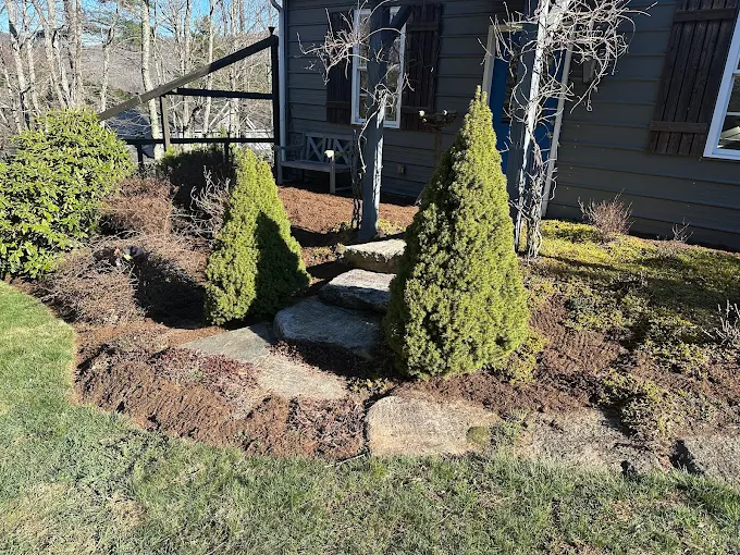 Natural stone slab steps with fresh dark mulch, trimmed evergreen shrubs, and garden beds in front of a home with gray siding and a blue front door.
